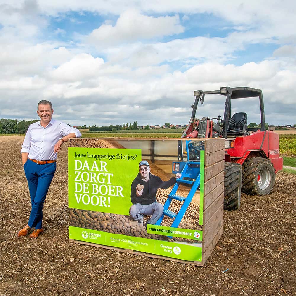 Op de achtergrond staat een tractor en op de voorgrond een man die op een kist met affiche leunt. Op de affiche staat een foto van een man bij een berg aardappels en de tekst "Jouw knapperige frietjes? Daar zorgt de boer voor!"